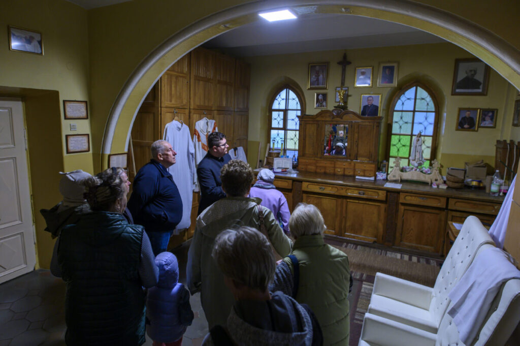 Grupa ludzi w pomieszczeniu kościelnym z drewnianymi meblami i kolorowymi witrażami. Na ścianie wiszą obrazy i krzyż. W tle widać figury religijne.