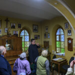 Grupa ludzi zwiedza pokój z witrażowymi oknami. Na ścianach wiszą zdjęcia i krzyż. Dominuje żółte oświetlenie i drewniane meble.