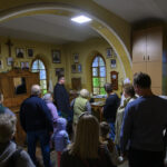 Grupa ludzi w pomieszczeniu z żółtymi ścianami i kolorowymi witrażami. Na ścianach wiszą zdjęcia. Arched okna dodają przytulności.