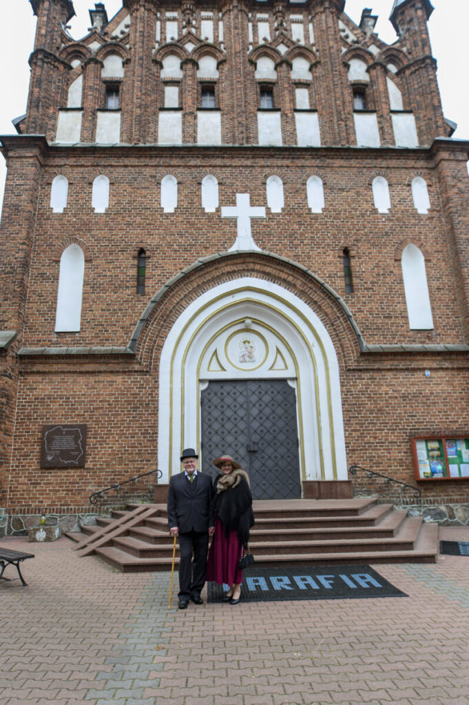 Para stoi przed zabytkowym, ceglastym kościołem. Widoczne schody i duże, zdobione drzwi. Strój elegancki, tło z czerwonej cegły.