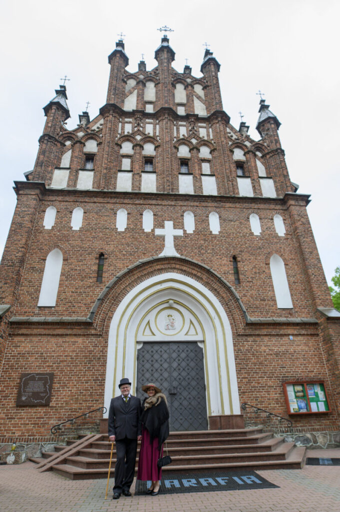 Ceglana fasada kościoła z licznymi ozdobnymi oknami. Przed wejściem stoi elegancka para. Na ziemi napis PARAFIA.
