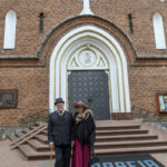 Para w eleganckich strojach stoi przed ceglastym kościołem. Drzwi są duże, ciemne, z białym łukiem i motywem świętego. Przed nimi schody i napis „PARAFIA”.