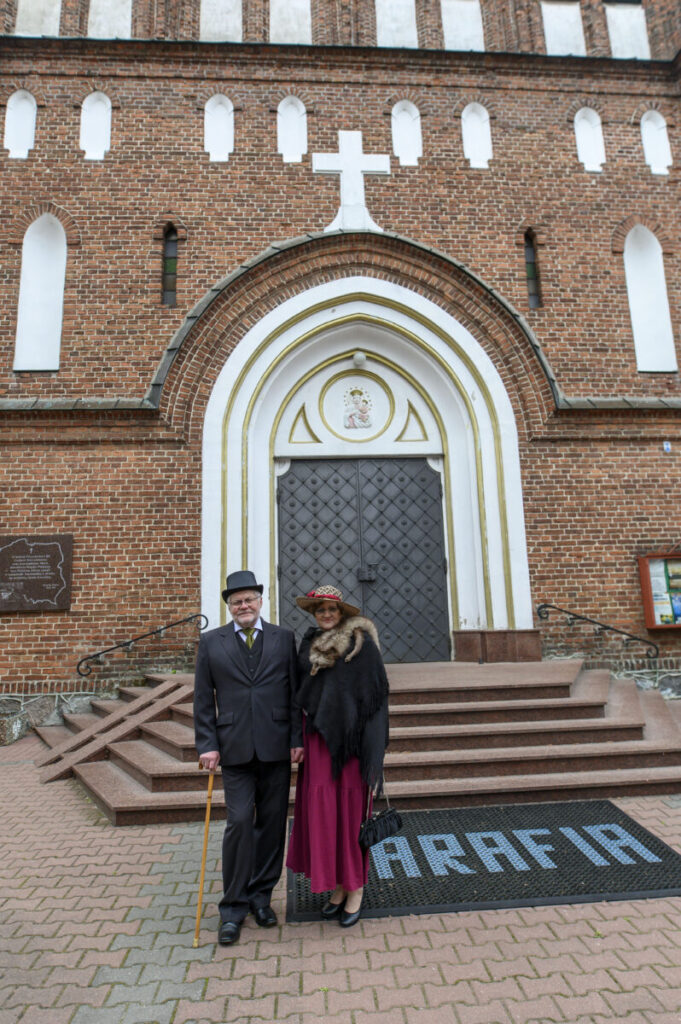 Para w eleganckich strojach stoi przed ceglastym kościołem. Drzwi są duże, ciemne, z białym łukiem i motywem świętego. Przed nimi schody i napis „PARAFIA”.