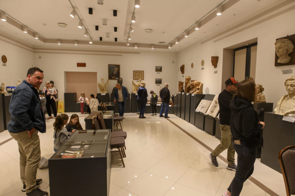 Wnętrze muzeum z rzeźbami na ciemnych podestach. Kilka osób ogląda eksponaty w jasnym pomieszczeniu. Sufit ozdobiony reflektorami.