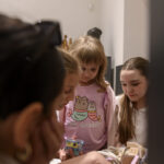 Grupa dzieci przegląda biały koszyk pełen przedmiotów. Jedno z dzieci ma różową bluzkę z grafiką. Tło jest jasne i neutralne.