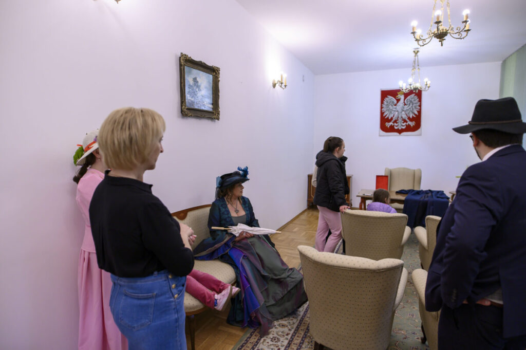Grupa ludzi w eleganckiej sali, niektórzy w kostiumach historycznych. Ściany białe z obrazem i godłem Polski. Oświetlenie lamp i żyrandola tworzy przyjemny nastrój.