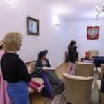 Grupa ludzi w eleganckiej sali, niektórzy w kostiumach historycznych. Ściany białe z obrazem i godłem Polski. Oświetlenie lamp i żyrandola tworzy przyjemny nastrój.