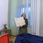 Osoba trzyma stary otwarty manuskrypt na tle zielonych żaluzji. Na stole obok wazon z białymi kwiatami. Przód zdjęcia to granatowa tkanina.