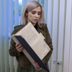 Kobieta trzyma otwartą teczkę z dokumentami. Ma jasne włosy, ubrana jest w ciemną kurtkę. W tle widać niebieskie żaluzje.