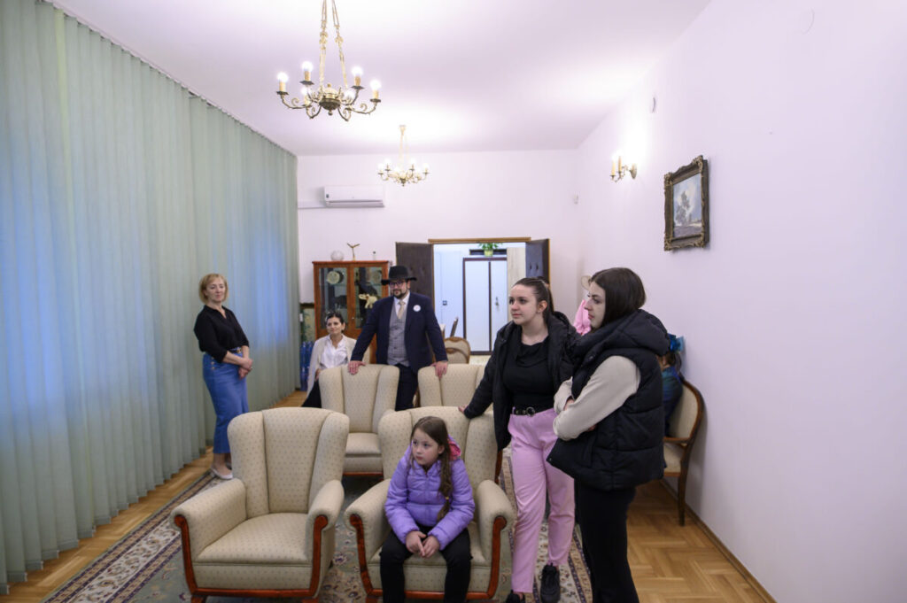 Pokój z jasnymi ścianami, trzema żyrandolami, zielonymi zasłonami. Ludzie siedzą i stoją wokół foteli i sofy. Na ścianie obraz.