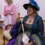 Dwie kobiety w kolorowych, stylowych strojach vintage siedzą i piją różowe wino. Jedna trzyma parasolkę. Tło jest jasne i neutralne.