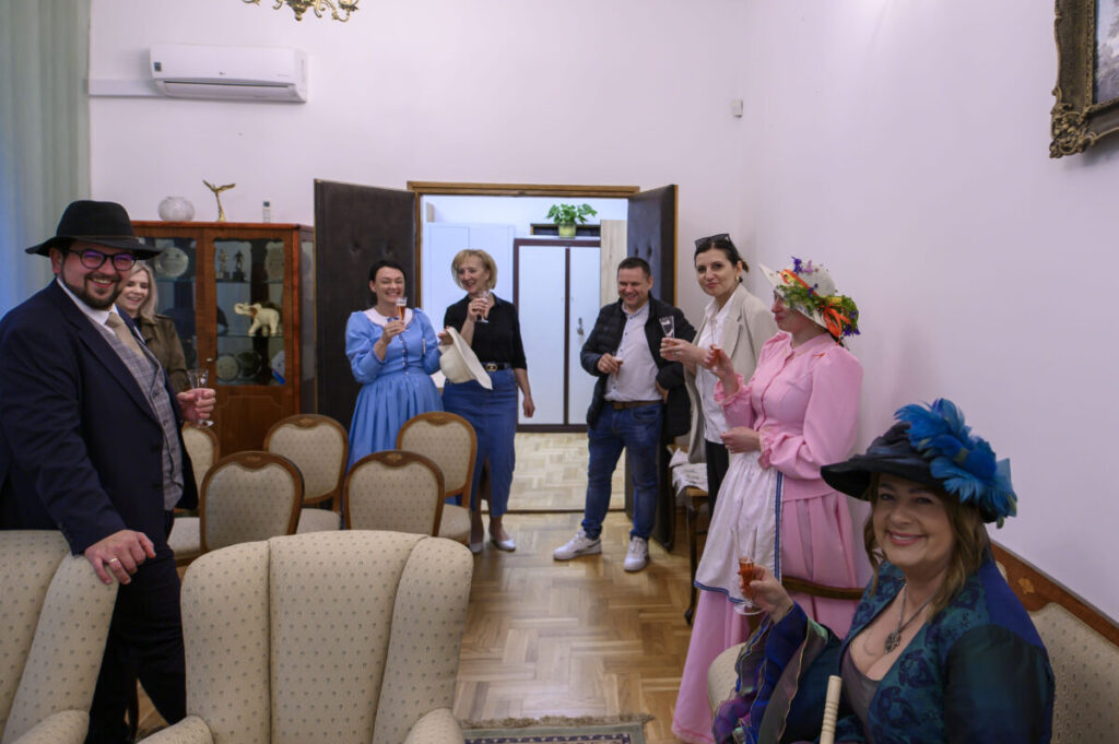 Grupa osób w eleganckich strojach stoi w salonie z jasnymi ścianami. W tle otwarte drzwi, widoczna szafka i rośliny. Ludzie trzymają kieliszki z napojem, uśmiechając się.