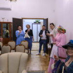 Grupa osób w eleganckich strojach stoi w salonie z jasnymi ścianami. W tle otwarte drzwi, widoczna szafka i rośliny. Ludzie trzymają kieliszki z napojem, uśmiechając się.