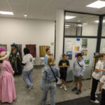 Grupa ludzi w kolorowych kostiumach i casualowych ubraniach na wystawie w galerii. Na ścianach wiszą kolorowe obrazy. Szare podłogi i białe ściany tworzą stonowane tło.