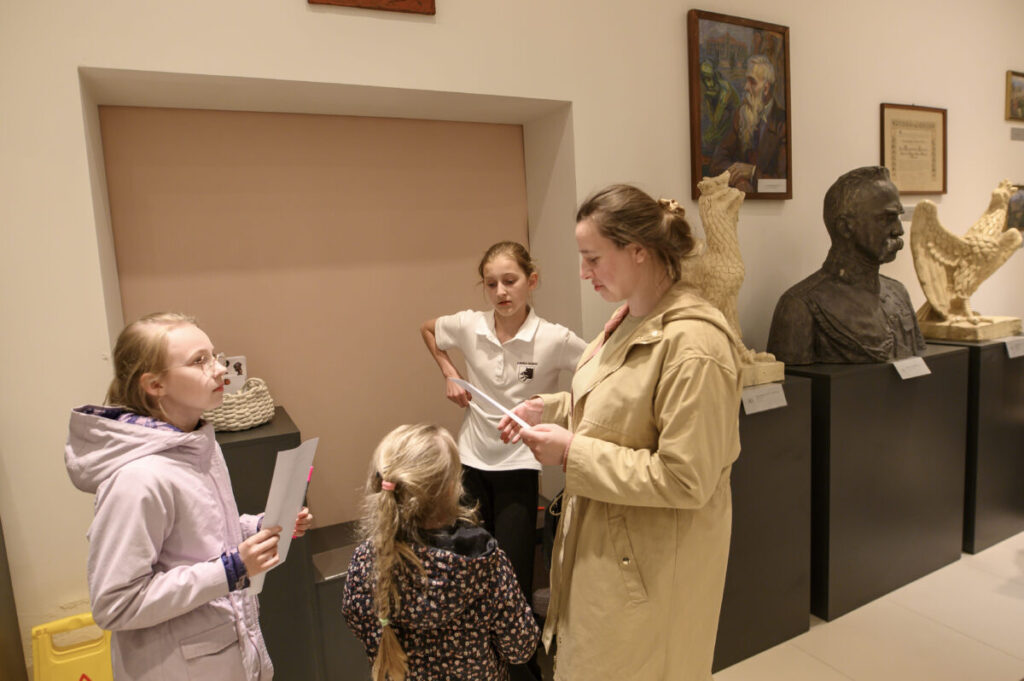 Cztery osoby rozmawiają w muzeum, otoczone rzeźbami i obrazami. Ściany są jasne, a osoby trzymają kartki. Pomieszczenie jest dobrze oświetlone.
