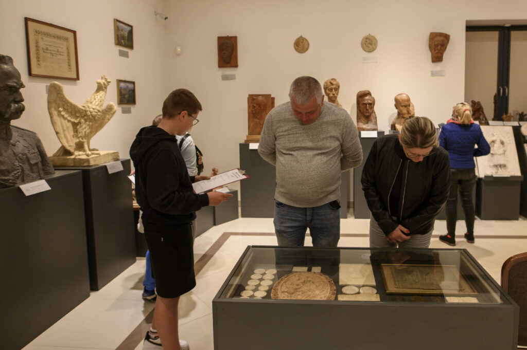 Mężczyzna i kobieta oglądają eksponaty w muzeum. W tle rzeźby i popiersia na ciemnych postumentach. Ściany jasne, podłoga beżowa.