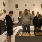 Mężczyzna i kobieta oglądają eksponaty w muzeum. W tle rzeźby i popiersia na ciemnych postumentach. Ściany jasne, podłoga beżowa.