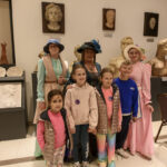 Grupa ludzi w muzeum, ubrani w historyczne stroje. Tło zdobią rzeźby i dekoracje kwiatowe. Uśmiechnięci, jasne kolory.