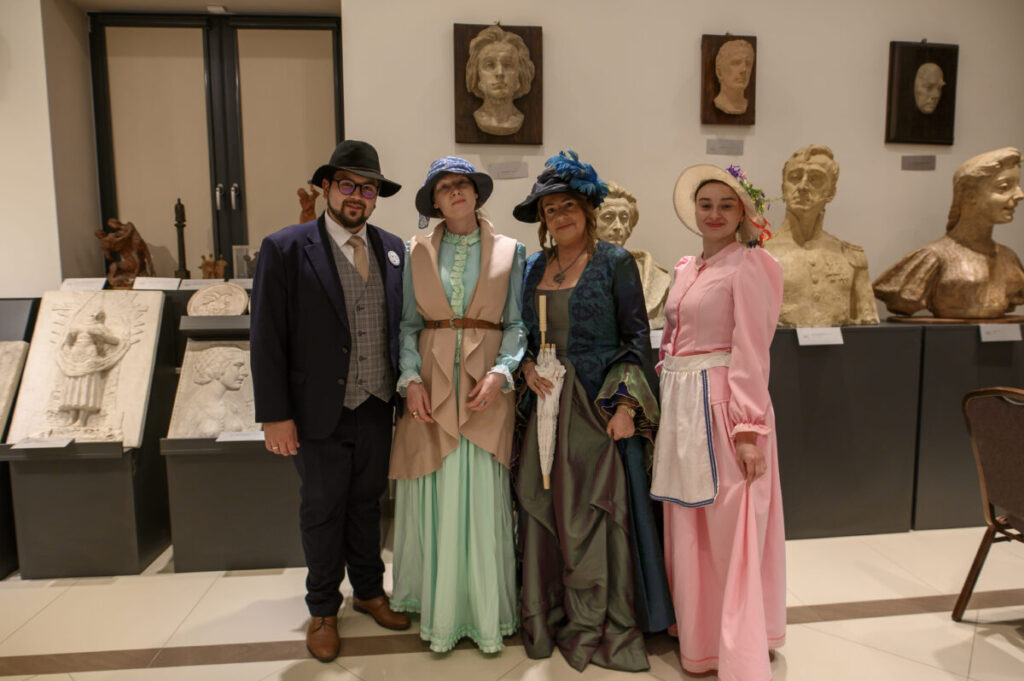 Cztery osoby w kostiumach retro pozują w muzeum. Tło to rzeźby i białe ściany. Kolory strojów to ciemny i pastelowy róż.