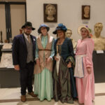 Cztery osoby w kostiumach retro pozują w muzeum. Tło to rzeźby i białe ściany. Kolory strojów to ciemny i pastelowy róż.