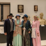 Cztery osoby w strojach retro pozują na tle rzeźb. Dominują kolory pastelowe i czarne. Tło jest jasne z popiersiami na ekspozycji.