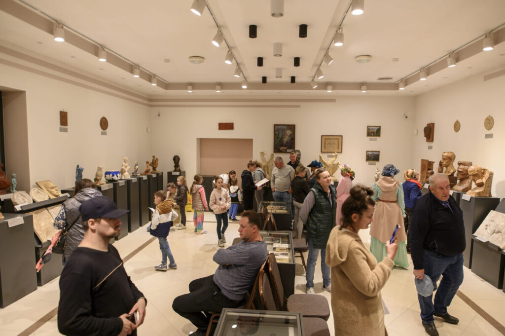 Sala galerii z białymi ścianami, rzeźbami na podestach, pełna zwiedzających. Jasne światło podkreśla detale eksponatów. Tło neutralne, podłoga w odcieniach beżu.