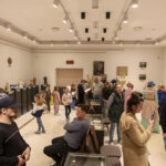 Sala galerii z białymi ścianami, rzeźbami na podestach, pełna zwiedzających. Jasne światło podkreśla detale eksponatów. Tło neutralne, podłoga w odcieniach beżu.