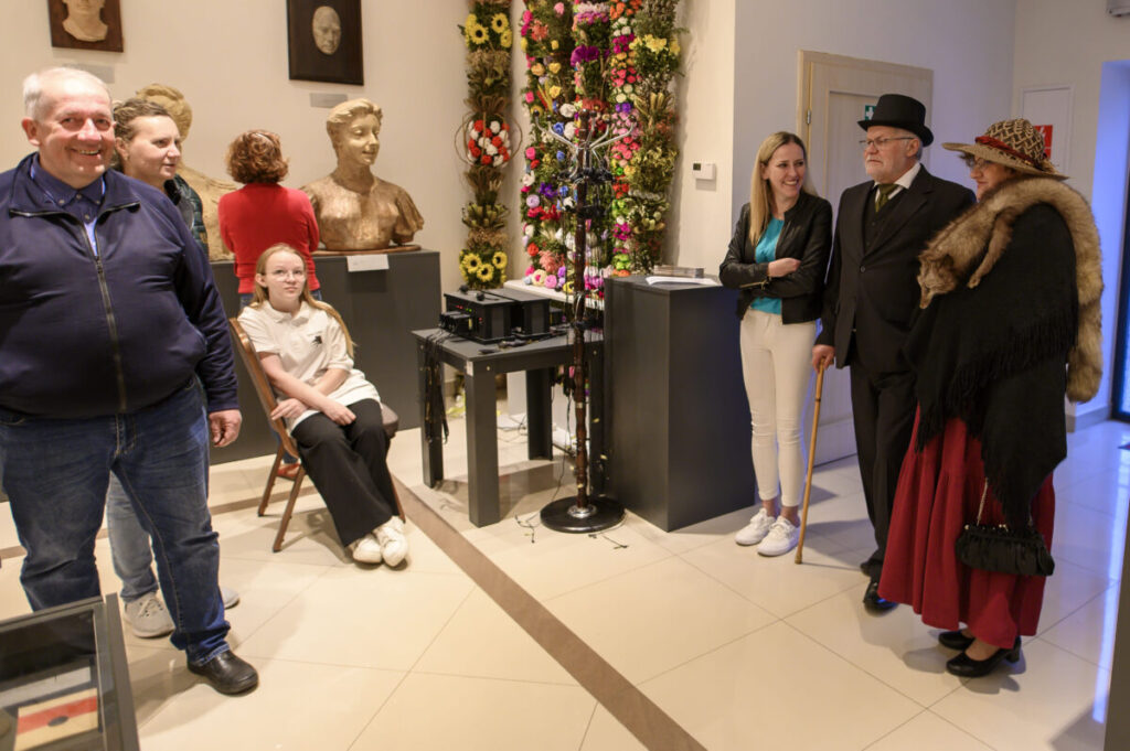 Grupa osób w muzeum, z rzeźbami i ścianą kwiatów w tle, jasne wnętrze. Mężczyzna w meloniku i kobieta z futrem.