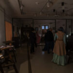 Pokój w stylu vintage, ciemno oświetlony. Ludzie w eleganckich strojach. Na ścianie wisi obraz, miedziane i beżowe tło.