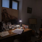 Biurko z fotografiami, biustem i maszyną do pisania. Na środku oświetlona lampka. W tle portret i pianino.