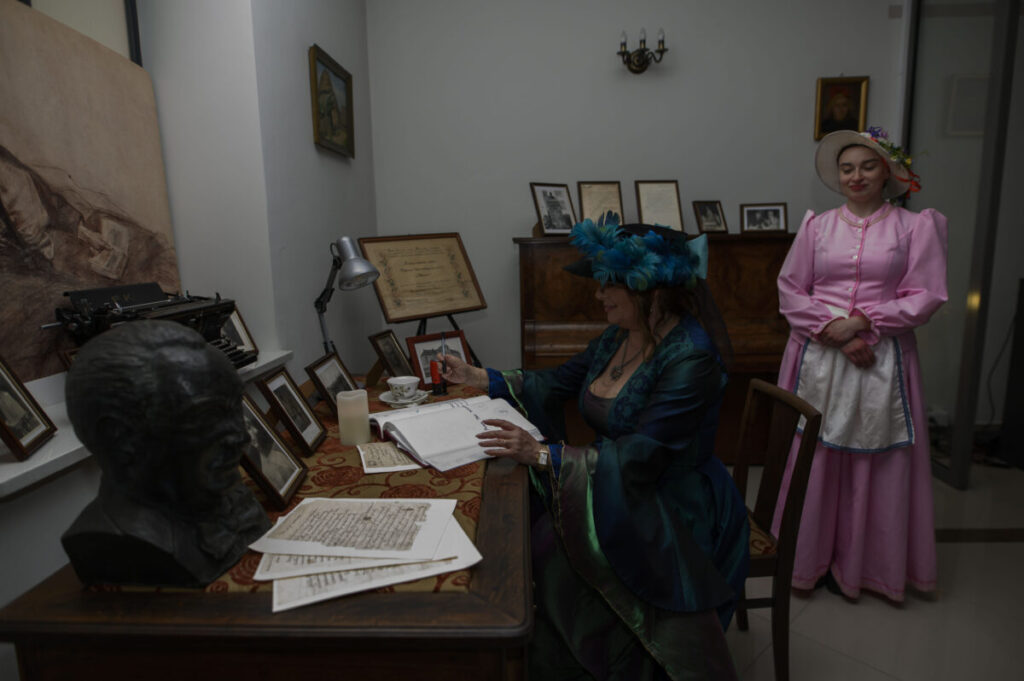 Na zdjęciu dwie kobiety w historycznych strojach siedzą i stoją w eleganckim pomieszczeniu z biurkiem. Kobieta siedząca pisze w księdze, ubrana w ciemnozieloną suknię z piórami na kapeluszu. Tło zdobi