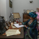 Kobieta w kapeluszu pisze w książce, obok stoi inna w różowej sukni. W tle biurko z popiersiem i dokumenty. Kolory ciemne i stonowane.