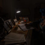 Kobieta w kapeluszu siedzi przy ciemnym stole, pisząc. Biurko oświetla mała lampka, wokół leżą ramki z zdjęciami. Na tle widoczna maszyna do pisania.