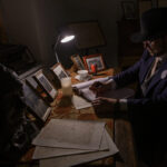 Mężczyzna w kapeluszu i okularach pisze przy biurku pod lampką. Na biurku zdjęcia, książki i kubek. Ciemne, nastrojowe tło.