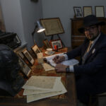 Mężczyzna w kapeluszu pisze przy biurku ozdobionym zdjęciami i biustem. Ściany zdobią dokumenty, a obok stoi maszyna do pisania. Pokój jest ciemny, oświetlony lampą.