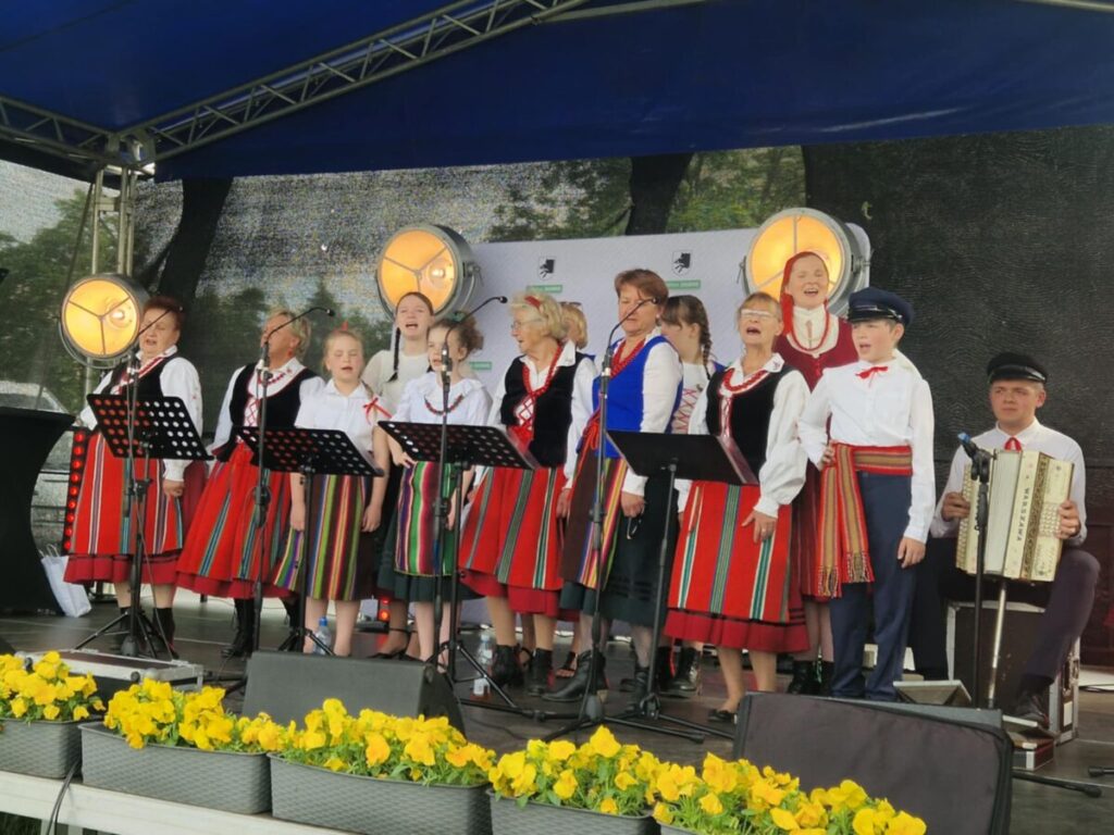 Grupa ludzi w strojach ludowych śpiewa na scenie. Mają czerwone i kolorowe spódnice oraz kamizelki. Tło zdobi niebieskie zadaszenie, a przód żółte kwiaty.