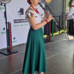 Osoba w ludowym stroju z zieloną spódnicą gra na klarnecie. Tło to jasny baner z logo Gmina Dobre. Jasne światła sceniczne oświetlają scenę.