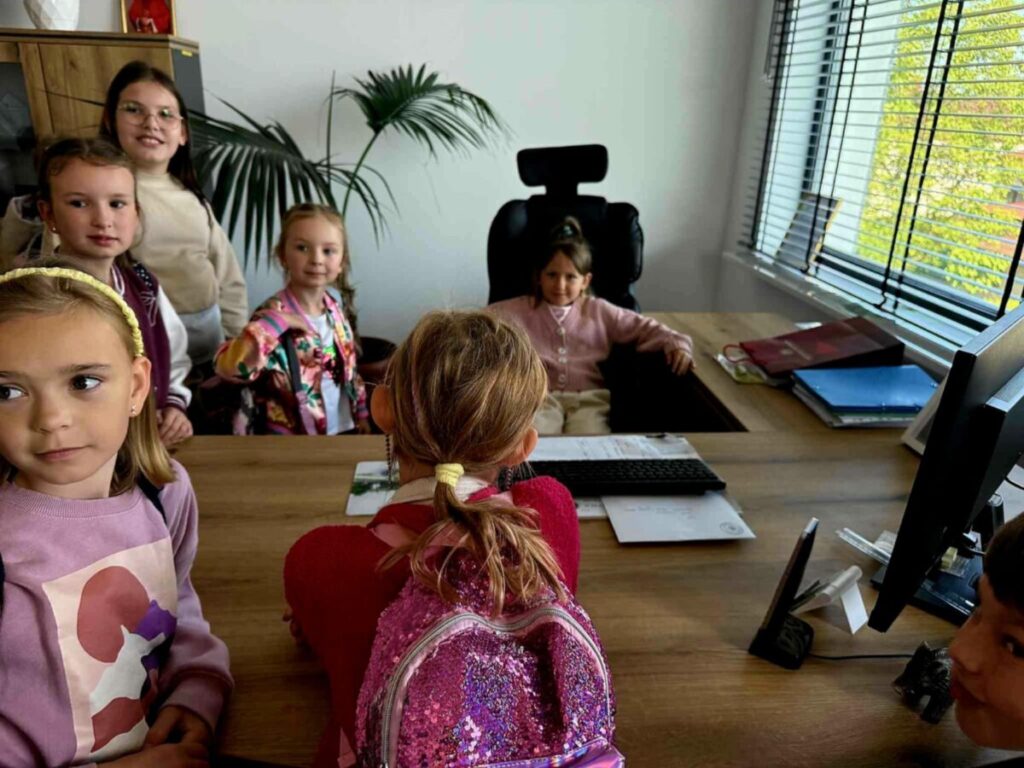 Grupa dzieci w kolorowych ubraniach stoi wokół biurka w nowoczesnym biurze. Jedno dziecko siedzi na czarnym fotelu, na biurku komputer i dokumenty. Przez okno z żaluzjami widać zielone drzewo.