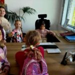 Grupa dzieci w kolorowych ubraniach stoi wokół biurka w nowoczesnym biurze. Jedno dziecko siedzi na czarnym fotelu, na biurku komputer i dokumenty. Przez okno z żaluzjami widać zielone drzewo.