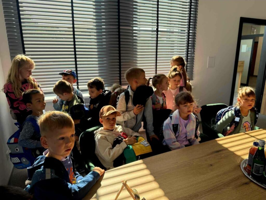 Grupa dzieci z plecakami siedzi w jasnym pokoju, ciepłe światło pada przez żaluzje. Pomieszczenie ma drewniany stół. Dzieci patrzą w stronę aparatu.