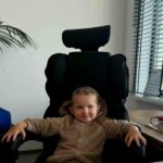 Małe dziecko w jasnym sweterku siedzi na czarnym fotelu biurowym. Tło to jasna ściana z rośliną i żaluzjami w oknach. Na biurku leżą dokumenty.