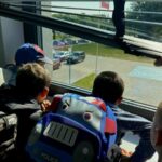 Dzieci z kolorowymi plecakami wyglądają przez okno w jasnym pomieszczeniu. Na zewnątrz widać parking i trawnik. Ręka dorosłego trzyma roletę.