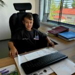 Młody chłopiec siedzi przy biurku w biurze, ubrany na ciemno. Przed nim klawiatura, wokół dokumenty. W tle duże okno z widokiem na dachy.