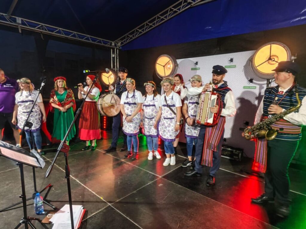 Ludzie w kolorowych, tradycyjnych strojach ludowych stoją na scenie. Jasne światła reflektorów oświetlają grupę. W tle ciemne niebo i białe tło.