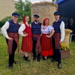 Pięć osób w tradycyjnych strojach ludowych stoi przed belą siana. Tło to zielona trawa i drzewa. Stroje mają żywe kolory: czerwony, biały i niebieski.