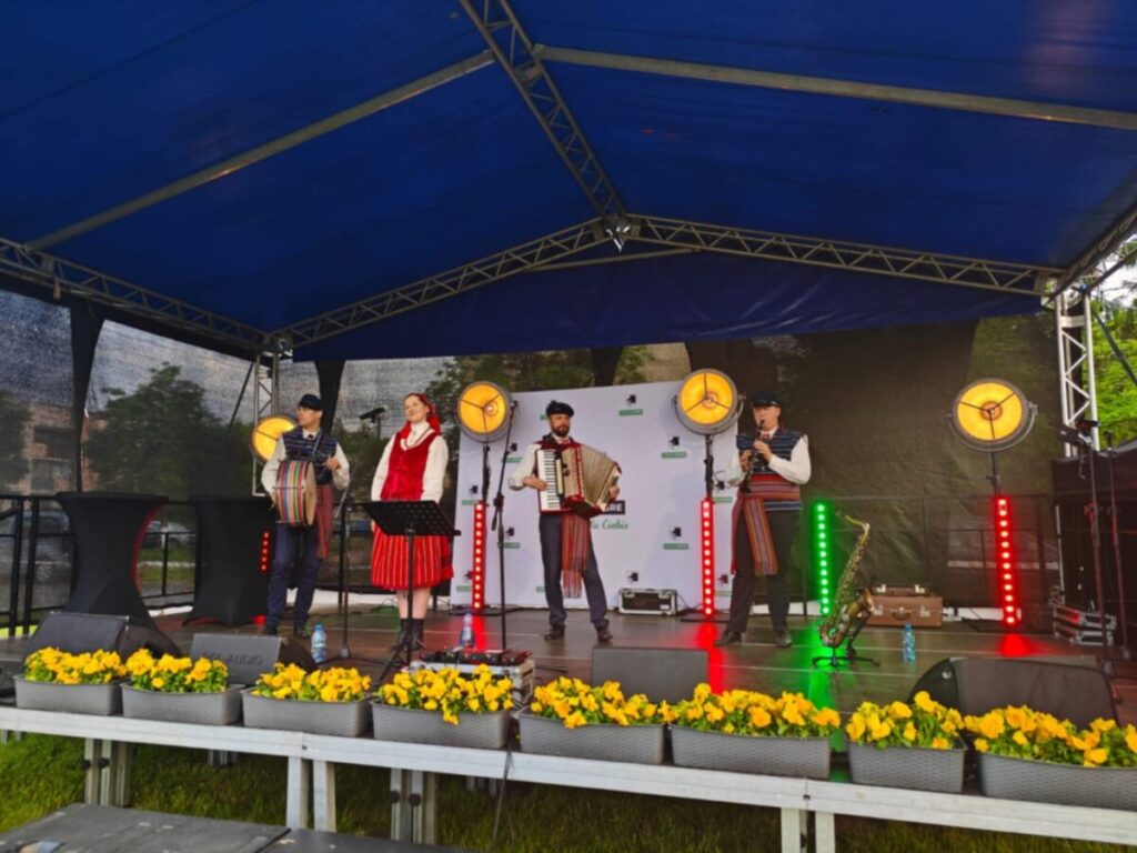 Grupa muzyków w tradycyjnych strojach na scenie pod niebieskim zadaszeniem. Tło z czarnym ogrodzeniem, scenę zdobią żółte kwiaty. Jasne reflektory oświetlają zespół.