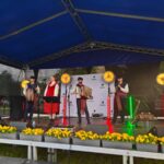 Grupa muzyków w tradycyjnych strojach na scenie pod niebieskim zadaszeniem. Tło z czarnym ogrodzeniem, scenę zdobią żółte kwiaty. Jasne reflektory oświetlają zespół.