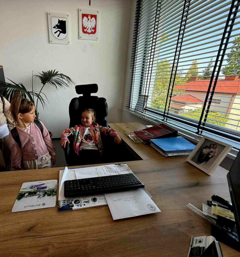 Dwoje dzieci w biurze; jedno siedzi na czarnym fotelu przy drewnianym biurku z klawiaturą. Biel ścian, godła, roślina i okno z żaluzjami. Na biurku zdjęcie w ramce i segregatory.