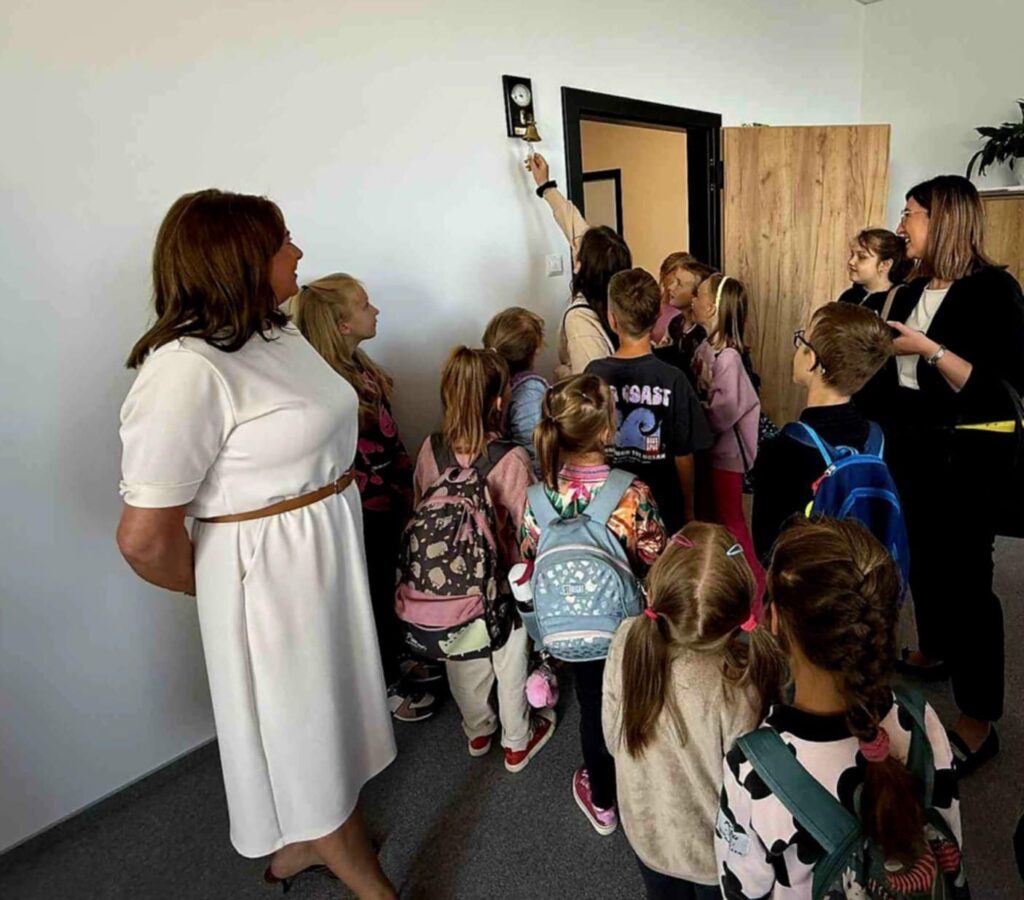 Grupa dzieci z plecakami stoi w pokoju z białymi ścianami, patrząc na kobietę dzwoniącą dzwonkiem. Obok dzieci stoją dwie kobiety. W tle znajdują się drewniane drzwi i roślina na górnej półce.