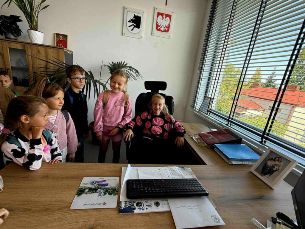 Grupa dzieci stoi w biurze przy biurku z klawiaturą; jedno dziecko siedzi na fotelu. Na ścianie są godła, a przez okno widać budynek z czerwonym dachem. Kolory są jasne i naturalne.